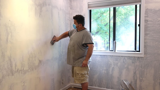 Man wearing working dust protective mask and sanding wall with a sanding block on preparation before