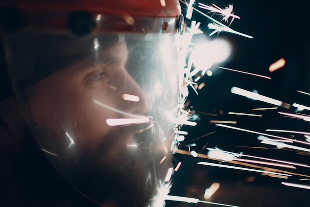 Photo man welder grinder in transparent protective mask with flying sparks in darkness.