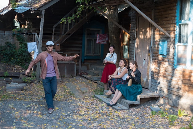 A man in white hat dancing russian folk dance women sitting on the stairs near the country house watching him and smiling a woman playing balalaika