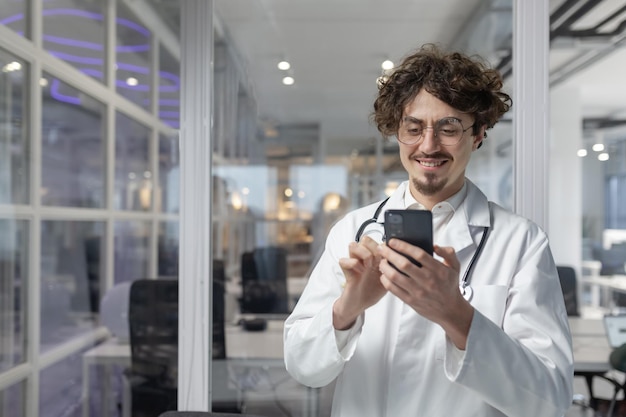 A man in a white lab coat and stethoscope holds a cell phone inside a medical office of a clinic