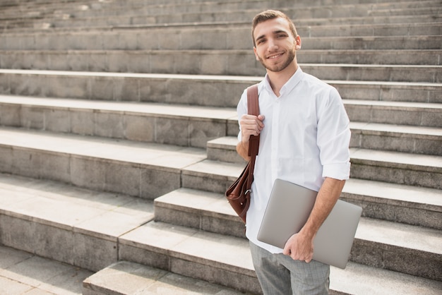 Man in white shirt holding a laptop and smiling at camera