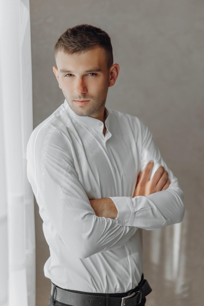 A man in a white shirt is standing in front of a window with his arms crossed