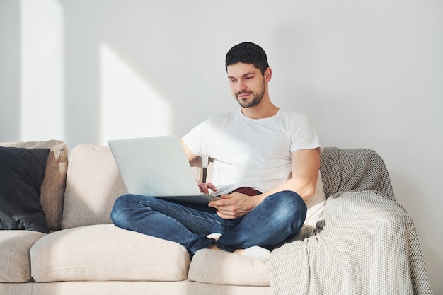 Man in white shirt and jeans sitting on bed with laptop indoors