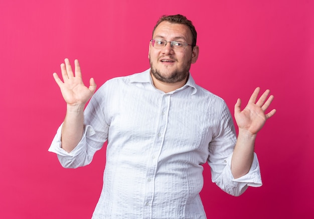 Man in white shirt wearing glasses  happy and positive raising arms standing over pink wall