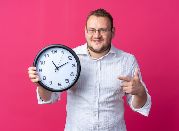 Man in white shirt wearing glasses holding wall clock pointing with index finger at it looking happy and cheerful smiling