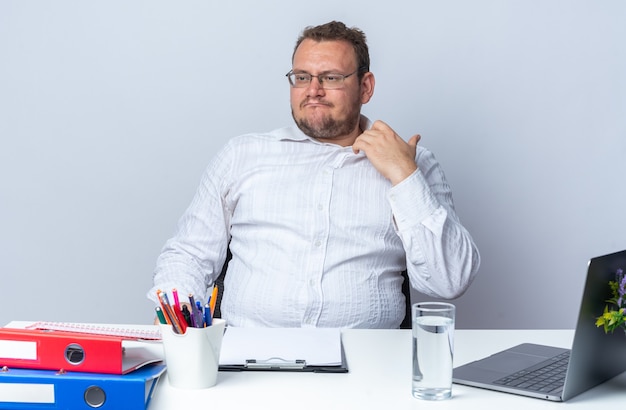 Man in white shirt wearing glasses looking aside annoyed and irritated touching his collar sitting at the table with laptop office folders and clipboard on white