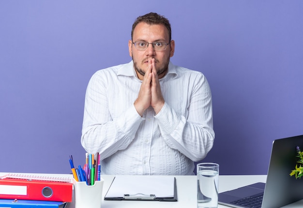 Man in white shirt wearing glasses with hands together stressed and nervous sitting at the table with laptop and office folders over blue wall working in office