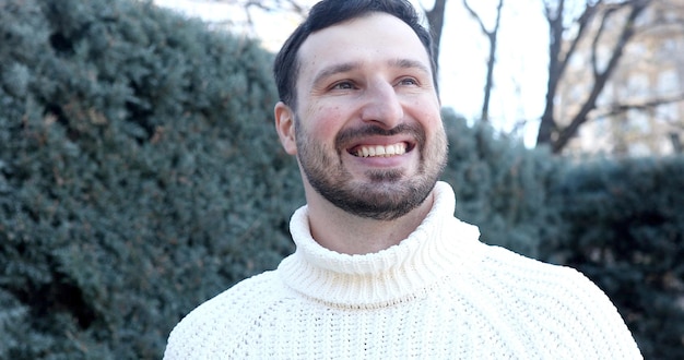 A man in a white sweater stands in front of a bush.