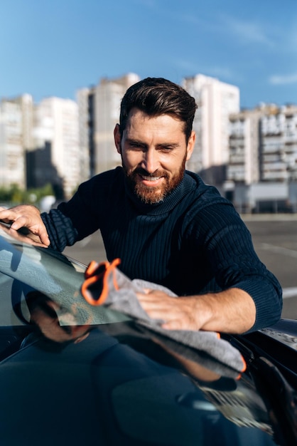 Man wiping his car at the street Car detailing wash during the sunny day Handsome bearded man in casual wear washing car doors and hood with microfiber cloth