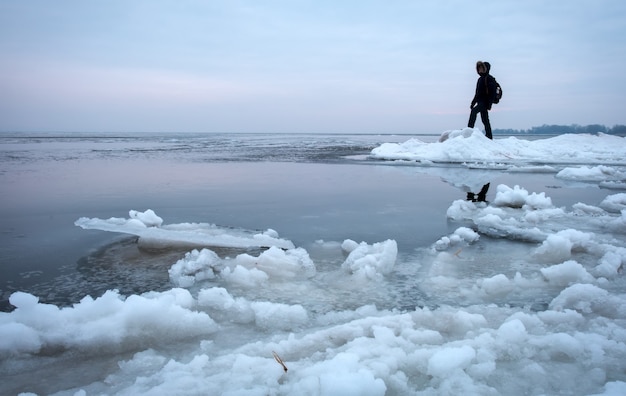 Photo man with backpack on the coast of frozen river during sunset. traveler concept