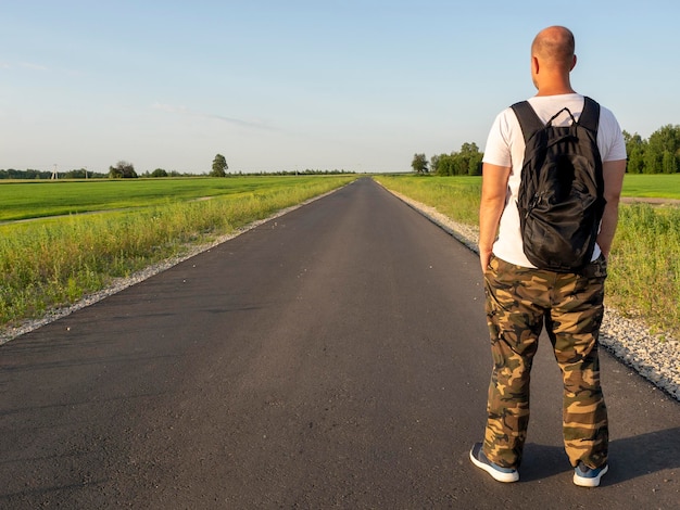 A man with a backpack on his back stands with his back to the camera on an empty asphalt road during the day The concept of travel and hiking adventure vacation Copy space