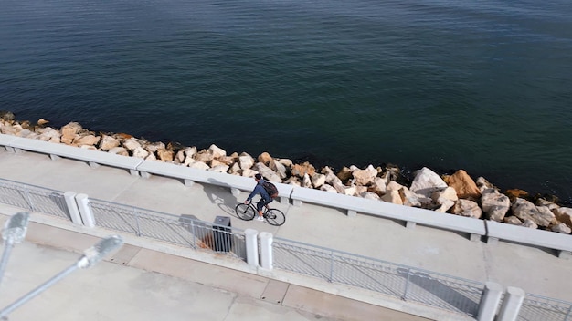 Photo a man with a backpack rides a bicycle along a seafront promenade seen from a flying drone