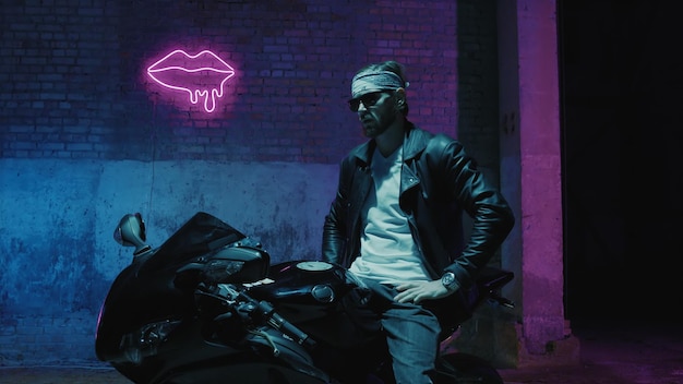 A man with a bandama sits on a sports motorcycle in sunglasses against a brick wall with a neon sign