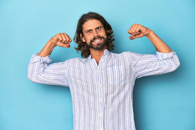 Photo man with beard in blue striped shirt blue studio showing strength gesture with arms