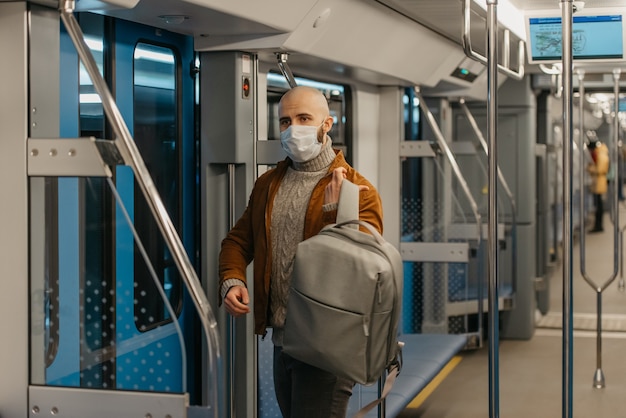 A man with a beard in a face mask to avoid the spread of coronavirus is putting on a backpack while riding a subway car. A bald guy in a surgical mask is keeping social distance on a train.