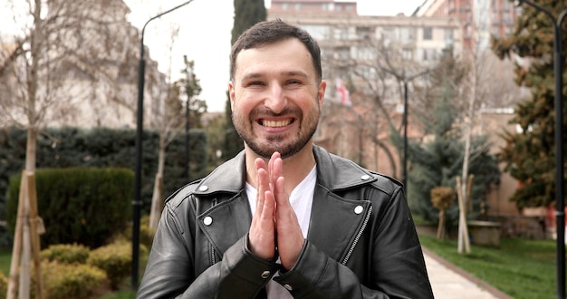 A man with a beard and mustache is standing in a park with his hands together.