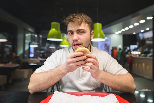 man with a beard sits in a fast-food restaurant with a burger in his hands