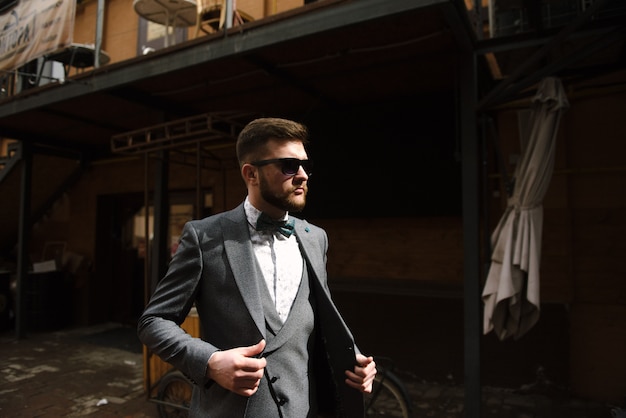 A man with a beard and sunglasses in a gray suit poses on the street to advertise men's clothing. 