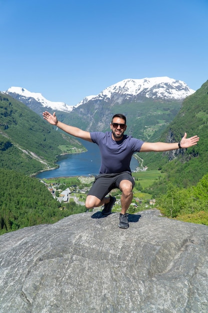 Man with beard and sunglasses raising his arms on a rock with the Geirangerfjord in the background and the snowcapped mountains on a sunny day