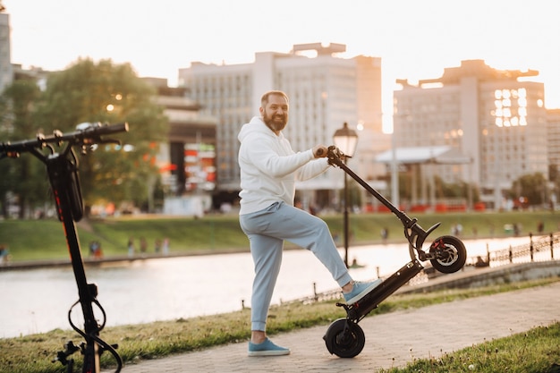 A man with a beard in white clothes rides around the city on an electric scooter at sunset.