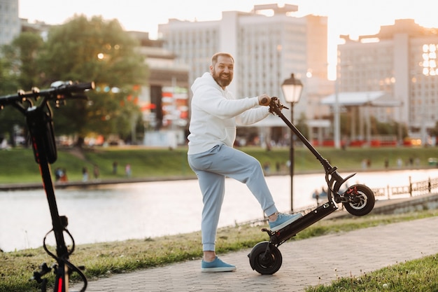 A man with a beard in white clothes rides around the city on an electric scooter at sunset.