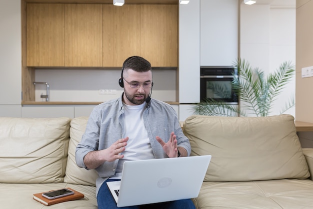 A man with a beard works from home sitting on the couch uses a laptop and a headset with a microphone conducts an online consultation