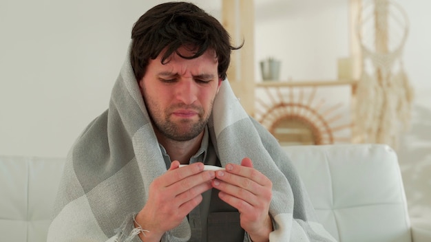 Man with a cold is sitting on the couch with a thermometer in his mouth