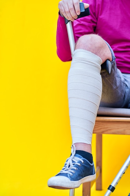 man with crutches, jeans and purple shirt sitting on a chair, supporting a bandaged leg on the crutch.