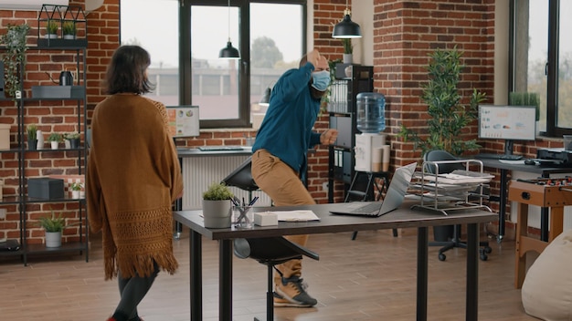 Man with face mask doing funny dance moves and celebrating victory after reading good news on laptop. Woman catching colleagues dancing and celebrating achievement in startup office.