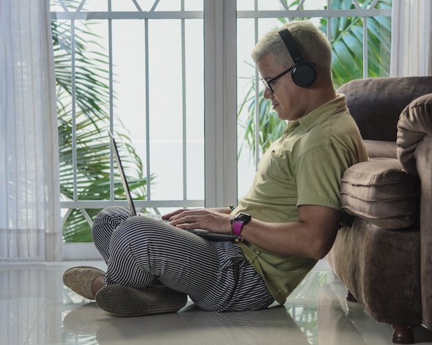 Man with glasses sitting on the living room floor leaning on a sofa next to a sale, checking laptop with headphones