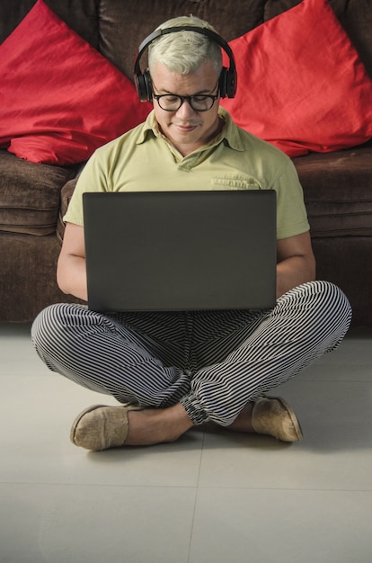 Man with glasses sitting on the living room floor next to the sofa, checking laptop with headphones