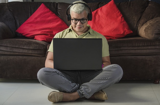 Man with glasses sitting on the living room floor next to the sofa, checking laptop with headphones