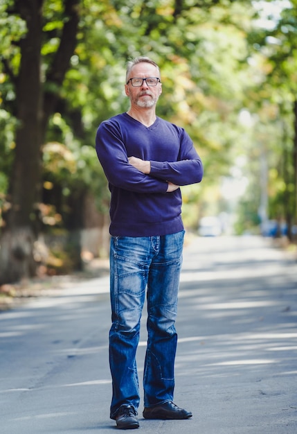 Man with gray hair outdoor in park No jacket Blue sweater Eyeglasses Bearded confident businessman Blurred background