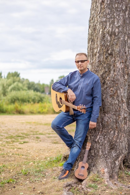 A man with a guitar leaning against a tree