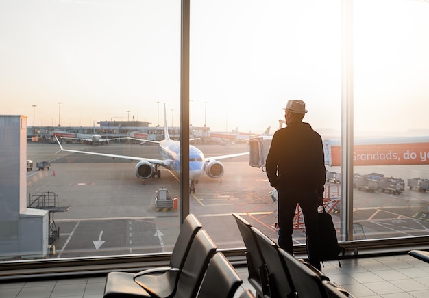 Man with hat and backpack is standing near window at the airport and watching plane