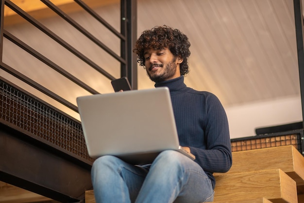 Man with laptop looking at smartphone sitting on stairs