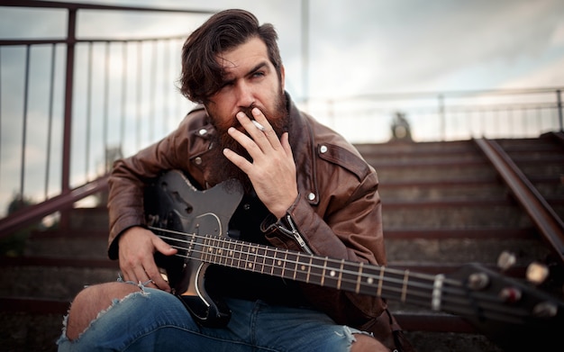 A man with a long beard in a brown leather jacket and torn jeans smokes a cigarette and holds an electric guitar while sitting on the steps