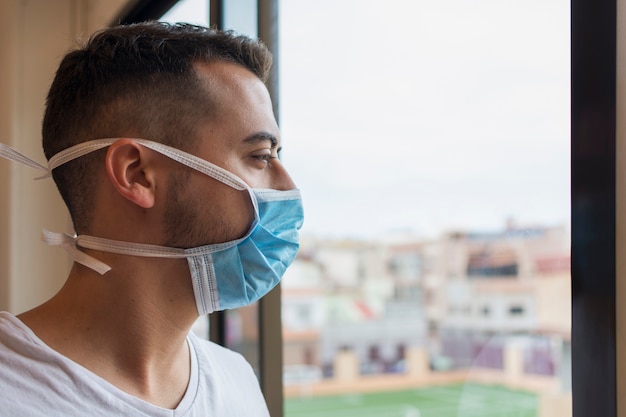 Man with a medicinal mask to protect himself against COVID-19, homebound, looking disconsolately through the window.