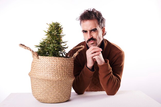 A man with a moustache stares at the camera next to a cannabis plant on a white background