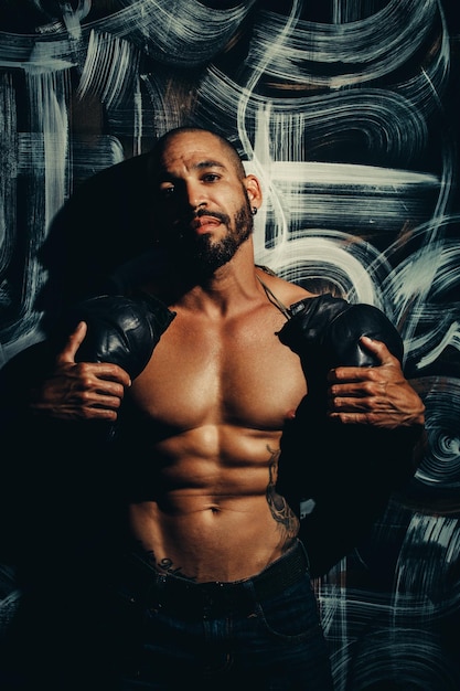 A man with a muscular body stands by the wall and holds boxing gloves A strong fighter with a naked torso