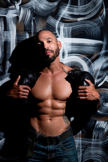 A man with a muscular body stands by the wall and holds boxing gloves A strong fighter with a naked torso