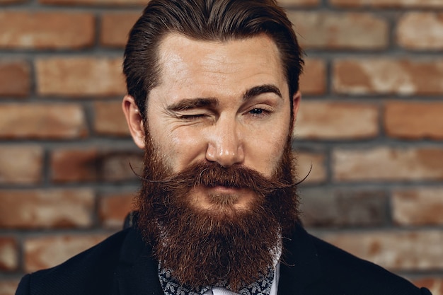 man with a mustache and beard posing against a brick wall and winking.