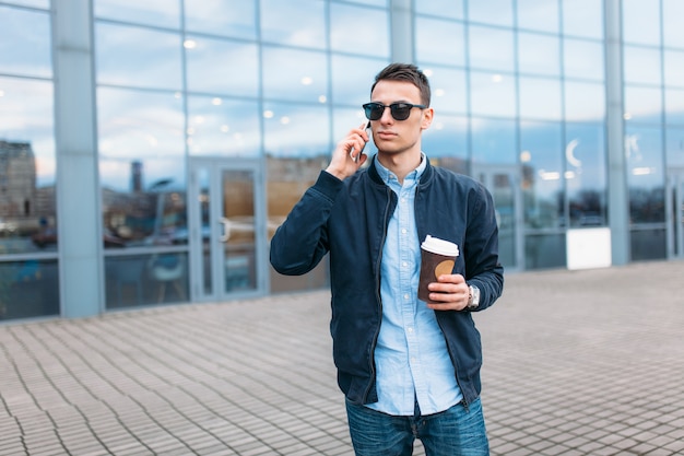 A man with a paper Cup of coffee, goes through the city, a handsome guy in stylish clothes and sunglasses, making a phone call