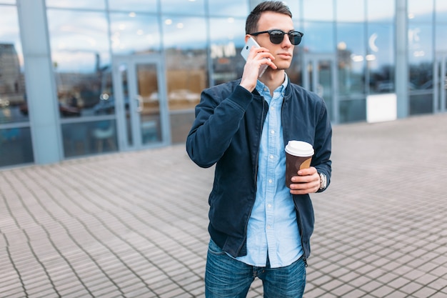 A man with a paper Cup of coffee, goes through the city, a handsome guy in stylish clothes and sunglasses, making a phone call