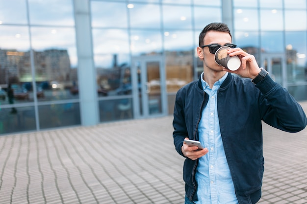 A man with a paper Cup of coffee, goes through the city, a handsome guy in stylish clothes and sunglasses, making a phone call