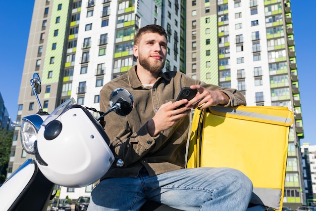 A man with a phone is looking for his order on the map Pizza delivery man on a scooter on the background of the city district