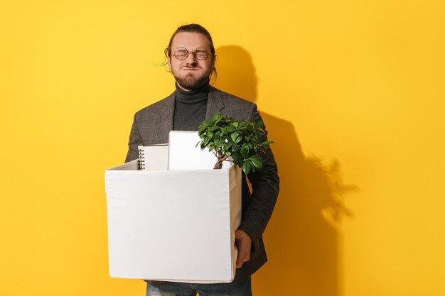 Man with silly face holding box with personal items after resignation against yellow background