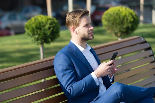 Man with smartphone in park. Manager with mobile phone on sunny outdoor. Businessman in blue suit sit on bench. Modern life and business lifestyle. Business communication, new technology and sms.