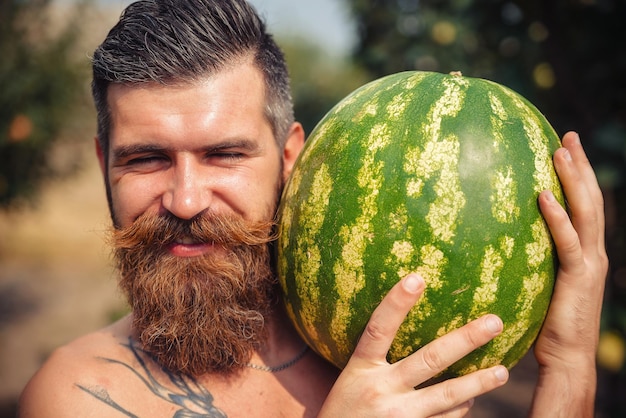 A man with a stylish hairstyle, a long beard and a tattoo keeps a big watermelon and smiles