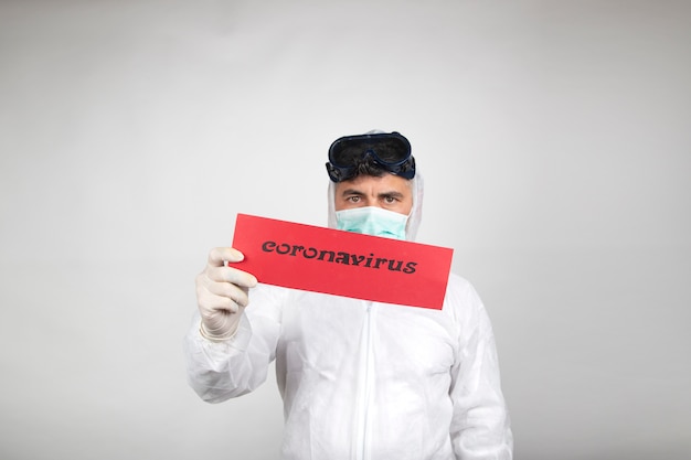 Man with surgical mask, glasses plus white protective suit and red sign with the word coronavirus isolated in studio on white background. New pandemic epidemic